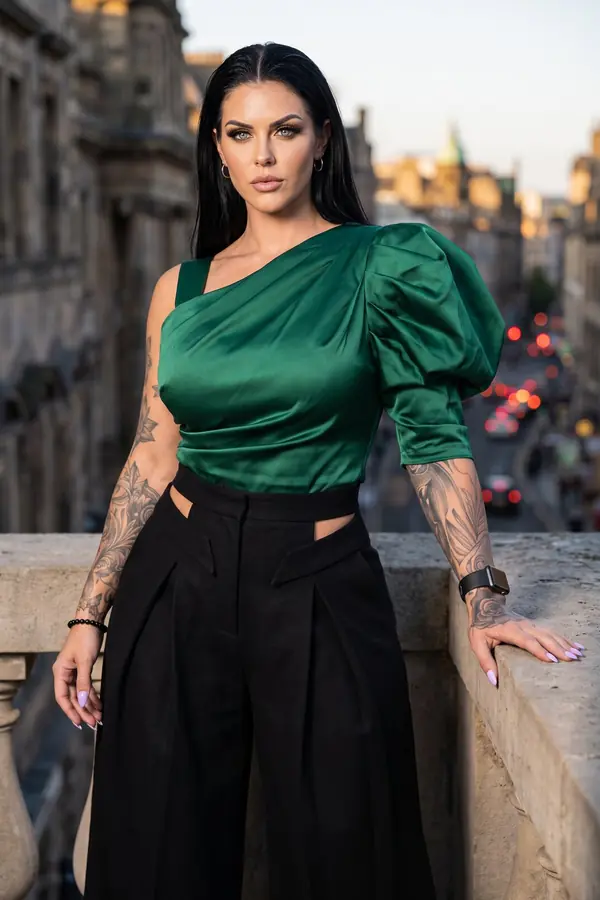 Woman in an emerald one-shoulder satin top and black pleated wide-leg trousers leaning on a stone balustrade at sunset