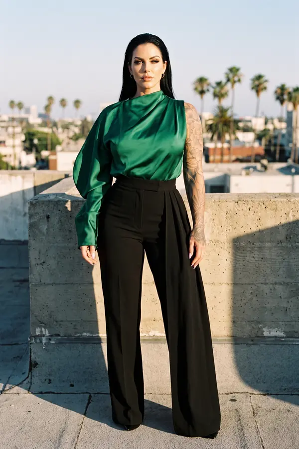 Woman in an emerald one-sleeve satin blouse and black pleated wide-leg trousers standing on a rooftop with palm trees behind