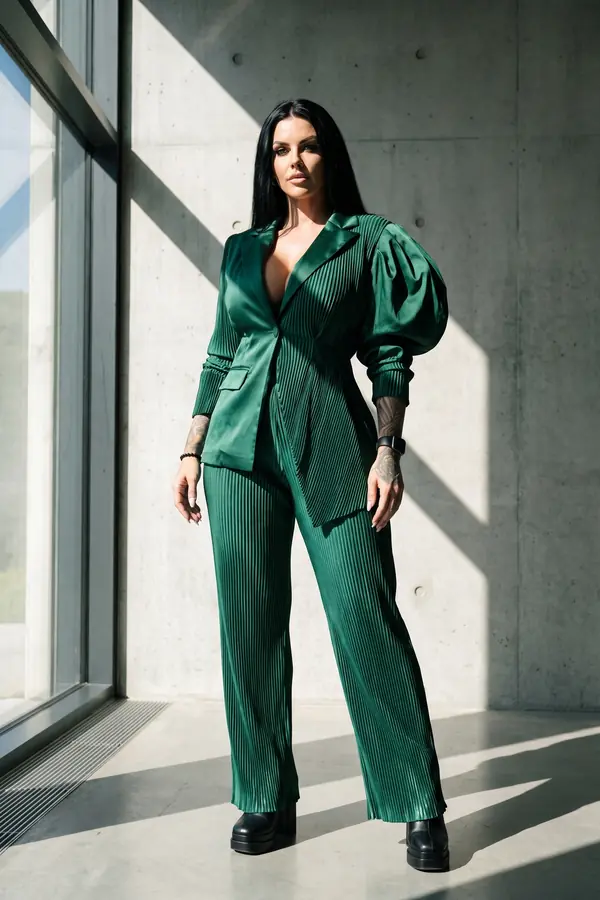 Woman in an emerald suit of smooth and pleated satin with one puffed sleeve, standing by a concrete wall