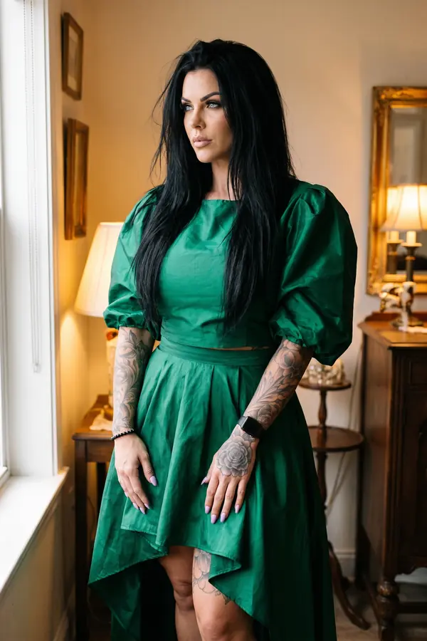 Woman in an emerald puff-sleeve crop top and matching high-low skirt standing by a window in a lamp-lit sitting room