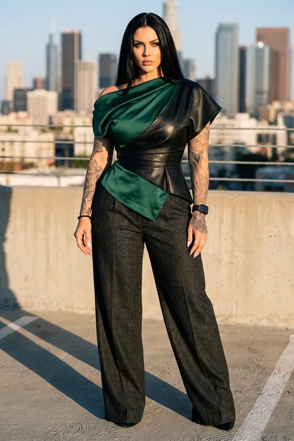 Woman in an emerald satin and black leather off-the-shoulder peplum top and charcoal wide-leg trousers on a rooftop