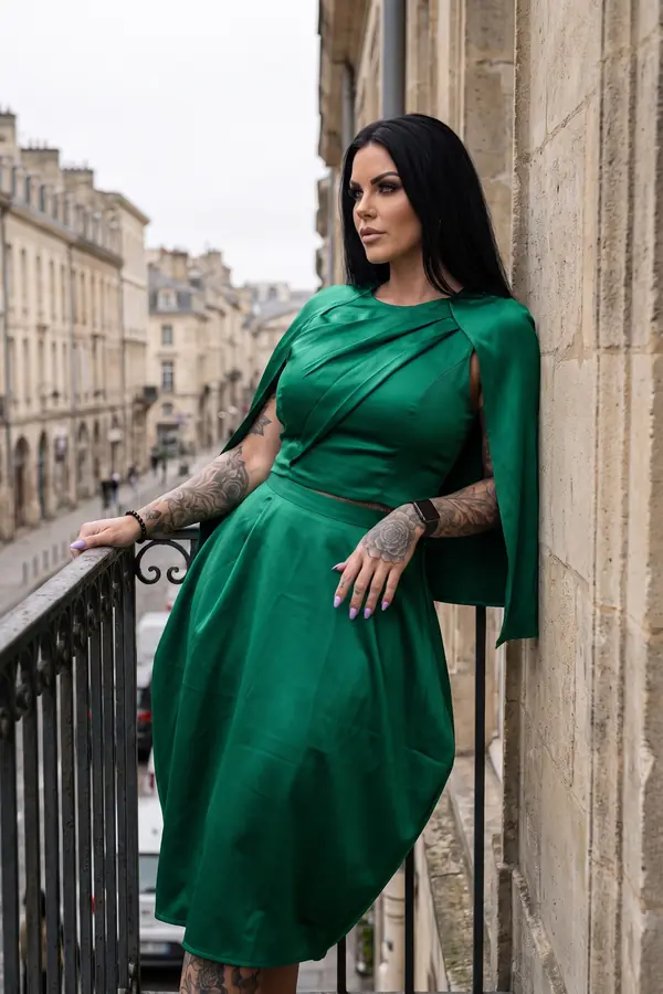 Woman in an emerald satin cape top and full midi skirt leaning against a stone wall on a city balcony