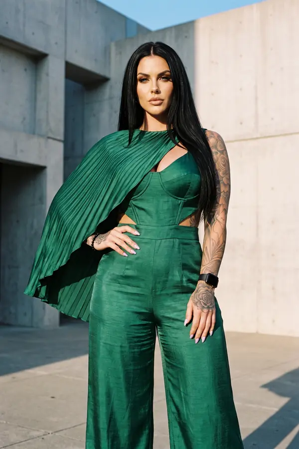 Woman in an emerald satin corset jumpsuit with a pleated cape sleeve standing in a concrete plaza in hard sunlight