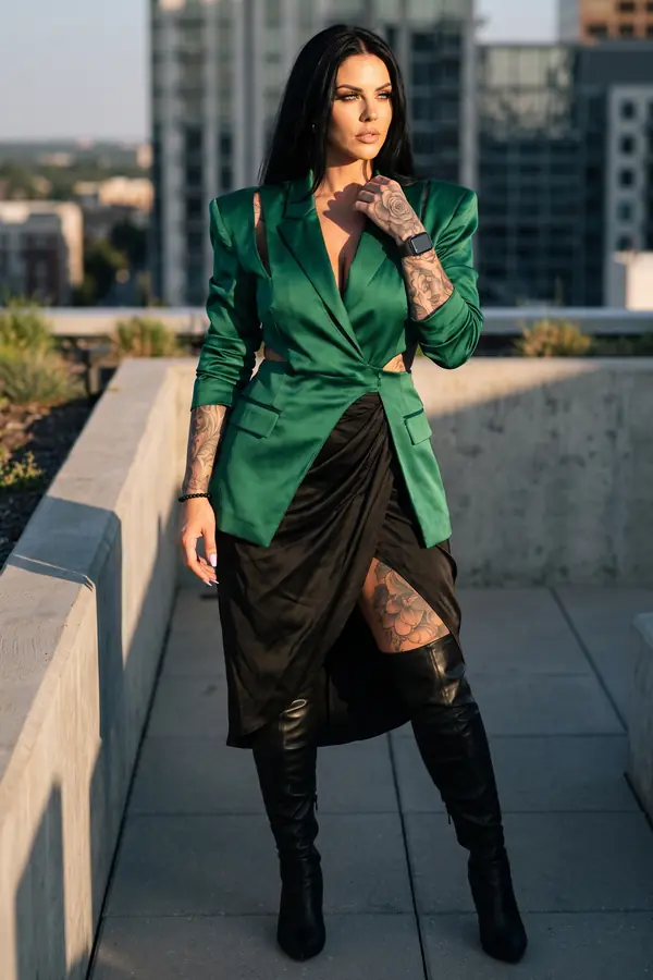 Woman in an emerald satin cut-out blazer, black draped satin skirt and over-the-knee leather boots on a rooftop at sunset