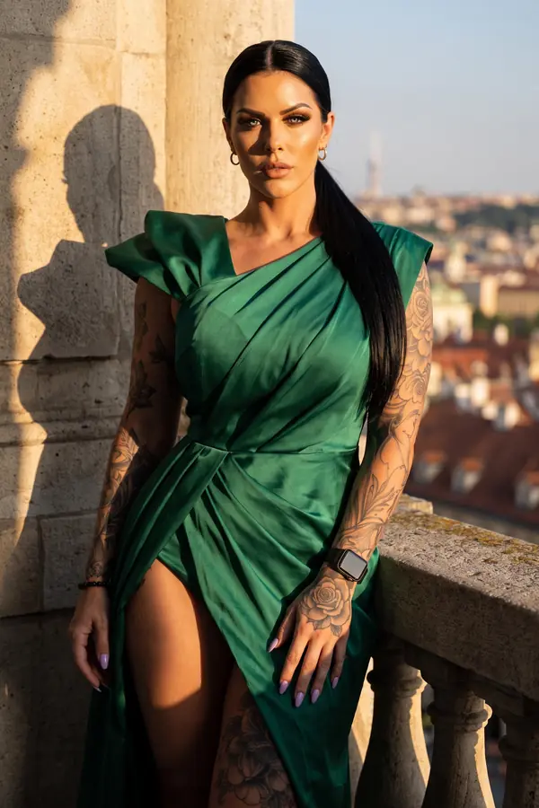 Woman in an emerald satin gown with padded shoulders and a side slit, leaning on a stone balustrade above a city at sunset