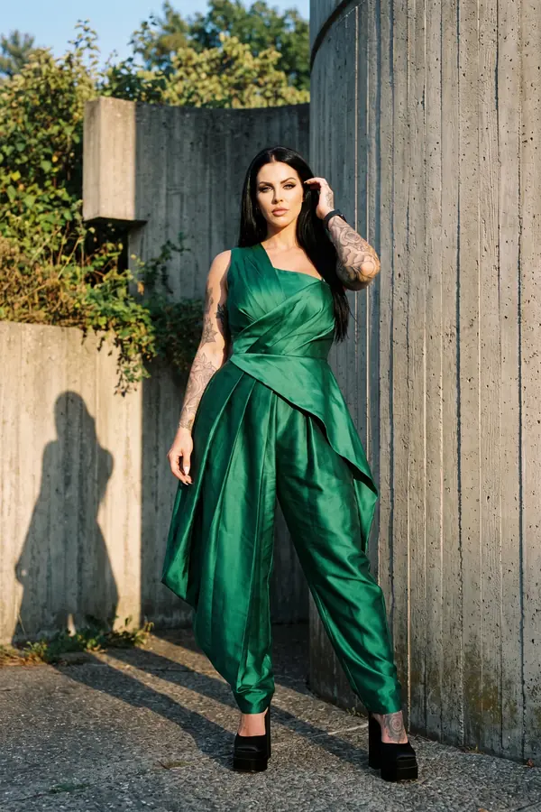 Woman in an emerald satin one-shoulder jumpsuit with draped hip panel and black platform heels beside a curved concrete wall