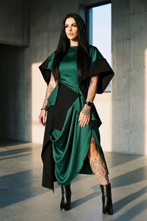 Woman in an emerald satin midi dress with black raw-edge panels and a side slit, in black ankle boots inside a concrete room