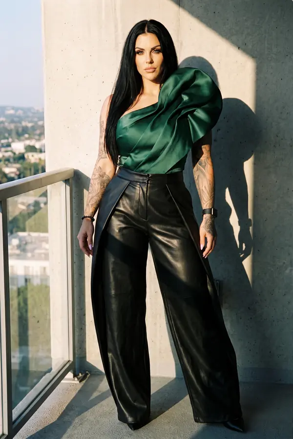 Woman in an emerald satin one-shoulder ruffle top and black leather wide-leg trousers on a high-rise balcony at sunset