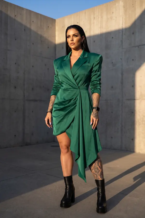 Woman in an emerald satin wrap blazer dress with an asymmetric drape and black platform boots in a concrete courtyard