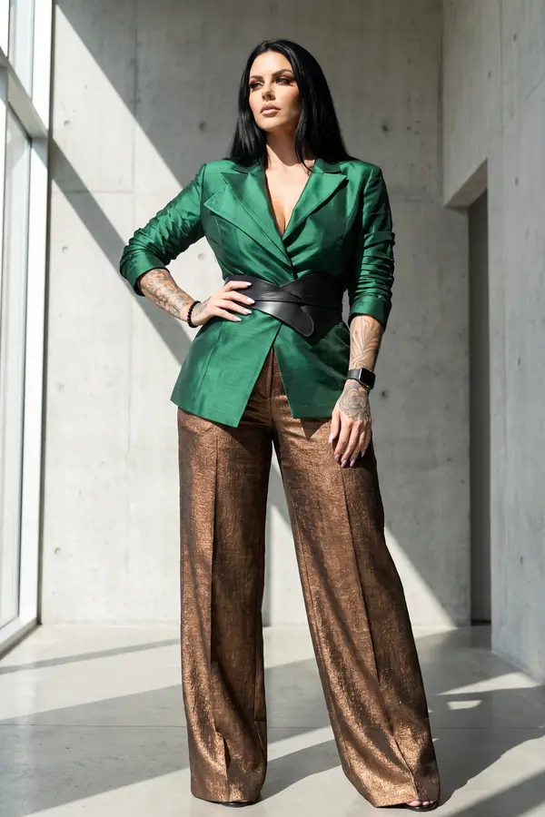 Woman in an emerald silk blazer with a wide black leather obi belt and bronze metallic wide-leg trousers in a concrete hall