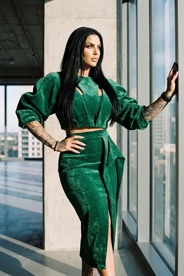 Woman in an emerald velvet puff-sleeve crop top and slit pencil skirt standing by a window on an empty office floor