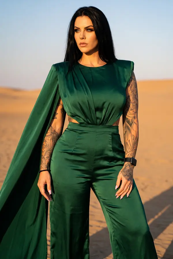Woman in a forest green satin jumpsuit with a long cape sleeve standing in desert dunes at golden hour