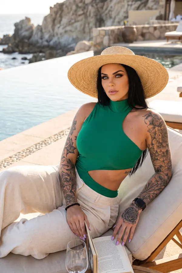 Woman in a green halter bodysuit, natural linen trousers and a straw hat on a lounger beside a pool above the sea