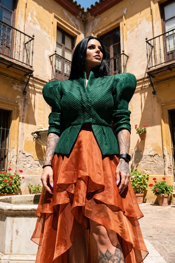 Woman in a green pleated puff-shoulder jacket and a tiered rust organza skirt in an old courtyard