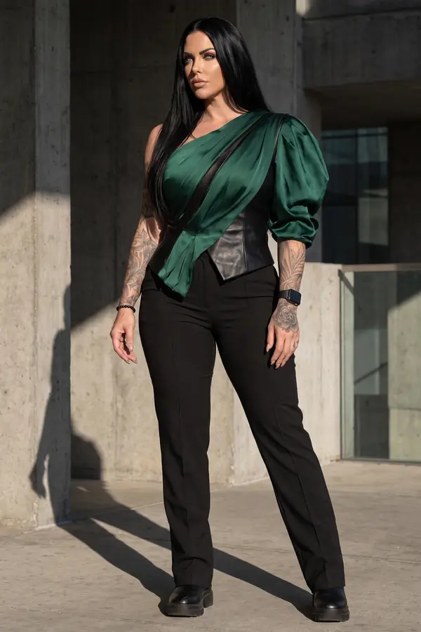 Woman in a green satin one-shoulder draped top over a black leather corset with black trousers by a concrete wall
