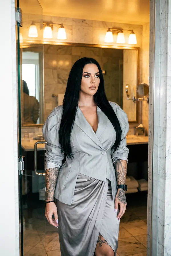 Woman in a pale grey wool wrap blazer and silver-grey satin draped skirt standing in a marble hotel bathroom doorway