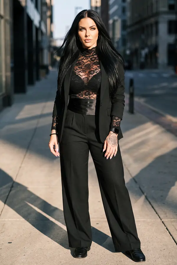 Woman in a high-neck black lace top under a black blazer with wide-leg trousers and a cummerbund on a downtown sidewalk