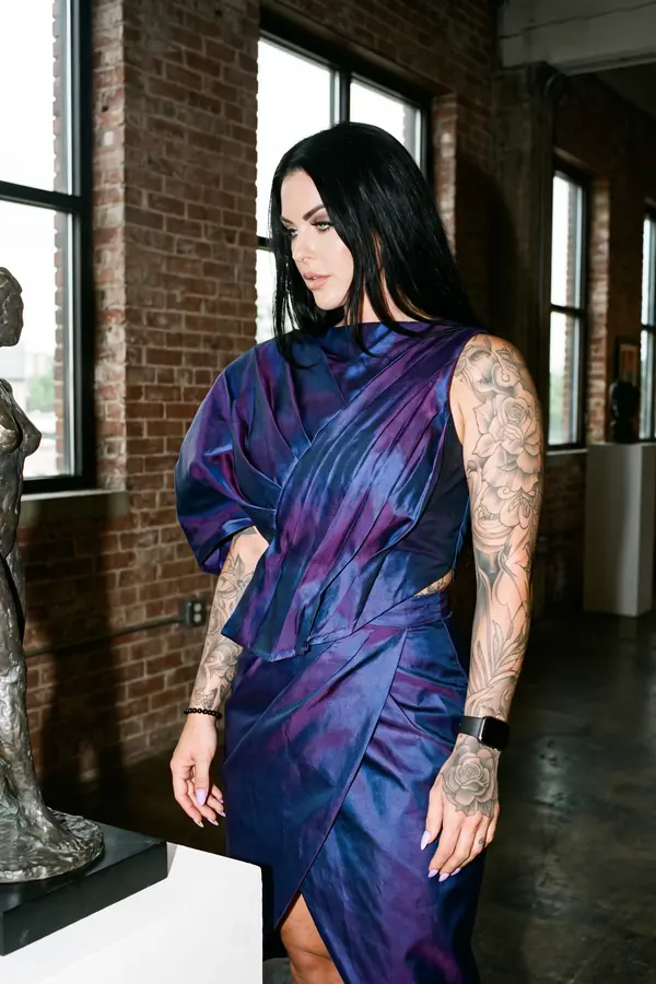 Woman in an iridescent navy and violet taffeta dress with one dolman sleeve beside a bronze sculpture in a gallery