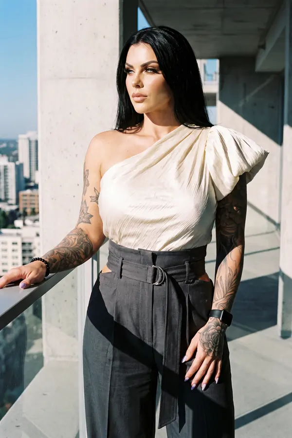 Woman in an ivory one-shoulder satin bow top and charcoal paperbag-waist trousers leaning on a high-rise balcony rail