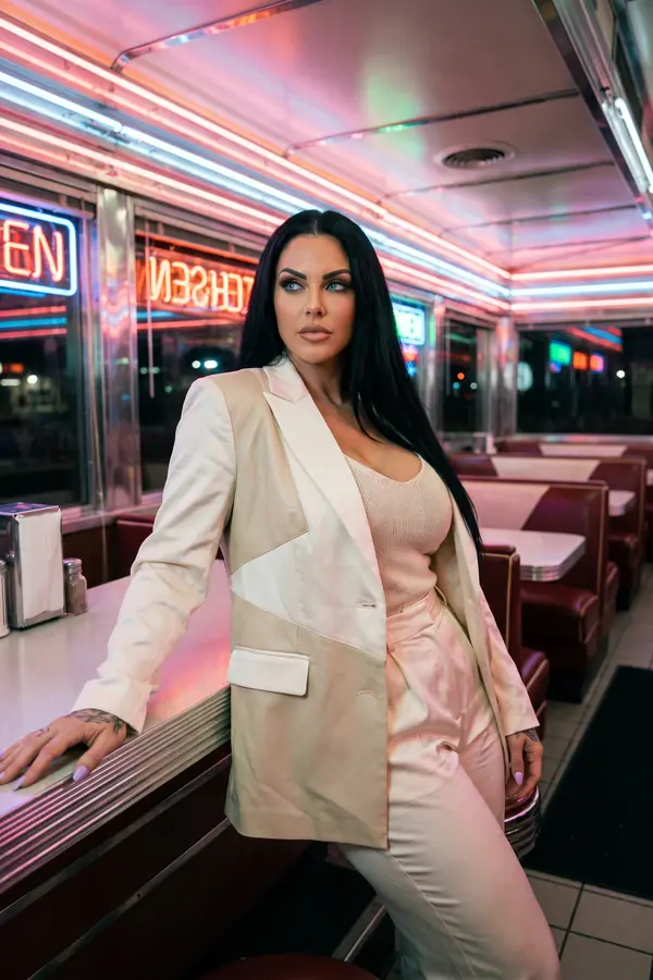 Woman in an ivory patchwork satin blazer, blush knit tank and cream trousers leaning on a diner counter under neon