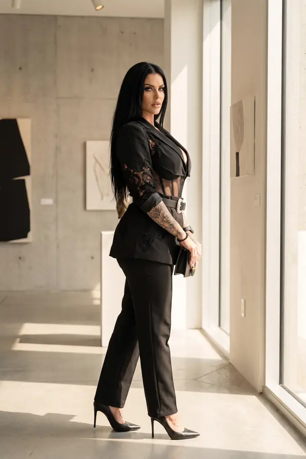 Woman in a black lace-inset blazer over a mesh corset with a crystal buckle belt, slim trousers and pumps in a gallery