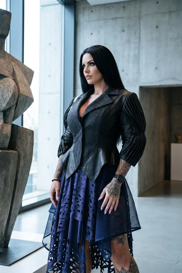 Woman in a charcoal wool and leather peplum jacket with pleated sleeves and a navy laser-cut skirt in a concrete gallery