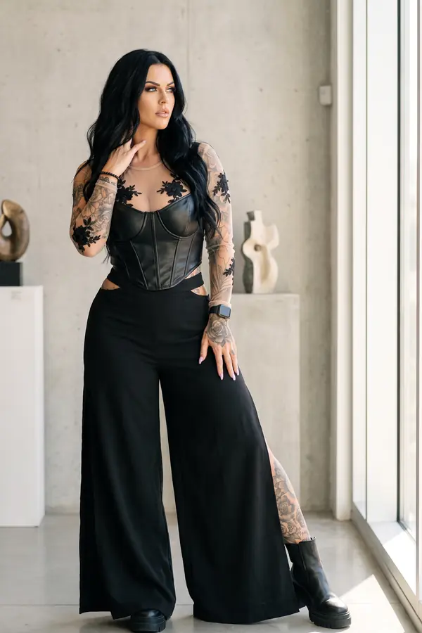 Woman in a black leather corset over a lace mesh top with black cut-out wide-leg trousers in a gallery