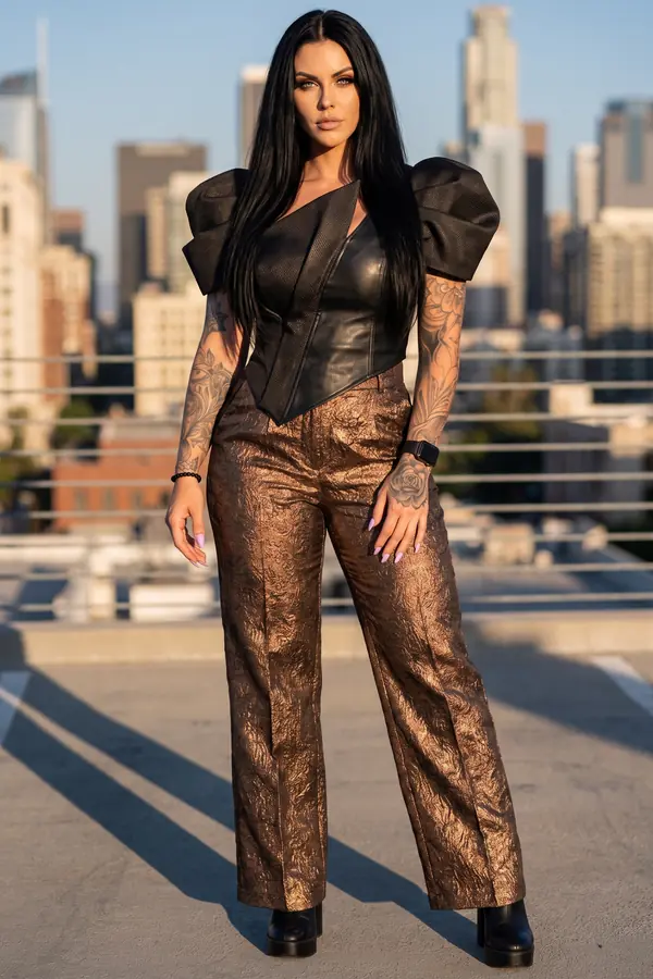 Woman in a black leather puff-sleeve top and bronze metallic brocade wide-leg trousers on a rooftop above a city skyline