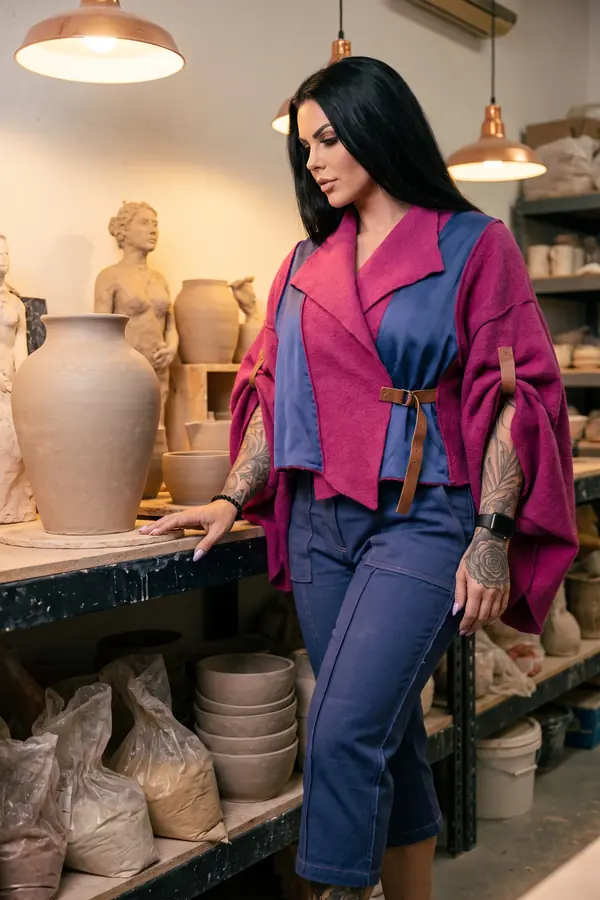Woman in a magenta wool crop jacket with blue satin panels and blue utility trousers in a pottery workshop