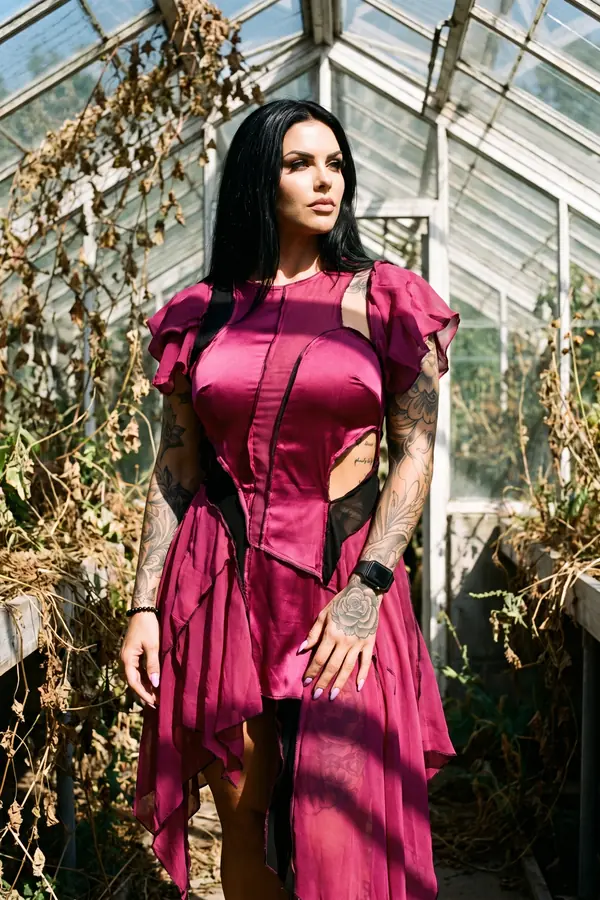 Woman in a magenta satin and chiffon dress with ruffle sleeves and black mesh panels in an overgrown glasshouse