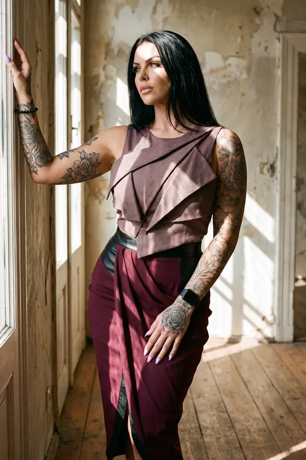 Woman in a mauve layered panel top and a burgundy draped skirt with a leather waistband, one hand on a window frame