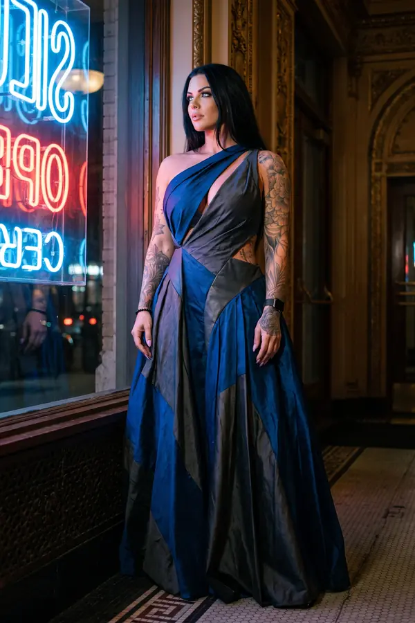 Woman in a one-shoulder navy and charcoal taffeta gown with leather inserts beside a neon shop window at night