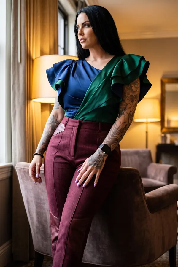 Woman in a navy and emerald satin ruffle-sleeve top and burgundy wool trousers leaning on a velvet armchair