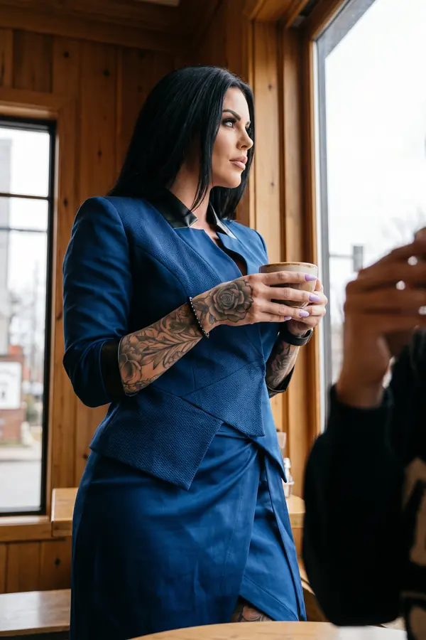 Woman holding a coffee cup by a window in a navy textured blazer with black leather collar and a navy wrap skirt