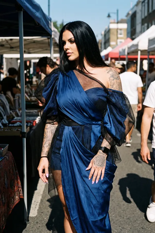 Woman in a navy draped satin one-shoulder dress with black mesh puff sleeves walking through a sunny street market