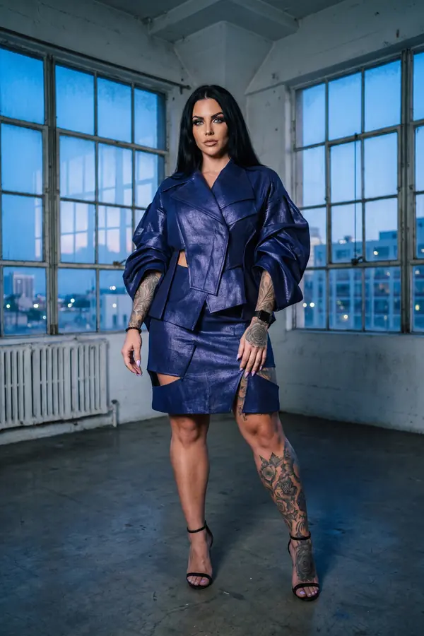 Woman in a shimmering navy deconstructed jacket and cut-out mini skirt with black sandals in an empty warehouse at dusk