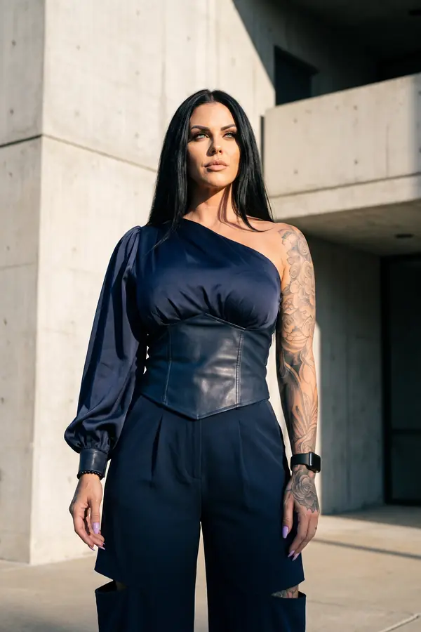 Woman in a navy satin one-shoulder corset-waist top and navy wide-leg trousers with leg cut-outs by a concrete building