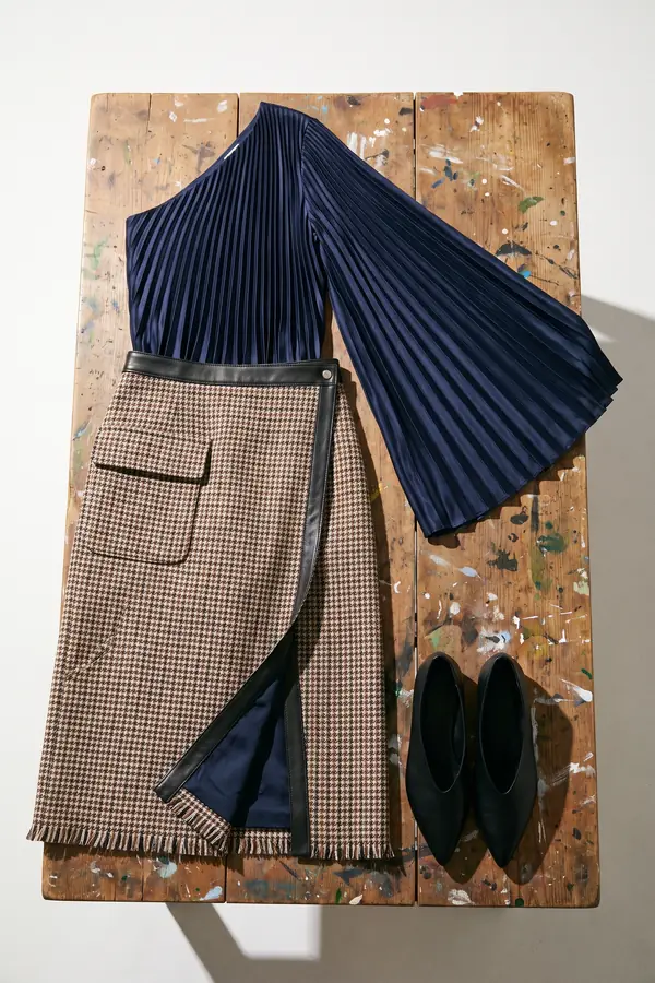 Woman painting at an easel in a navy pleated one-shoulder top and a brown check tweed wrap skirt with a fringed hem