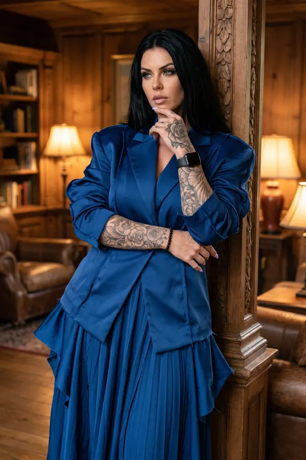 Woman with long black hair in an oversized navy raw-edge satin blazer and pleated maxi skirt leaning on a wooden pillar