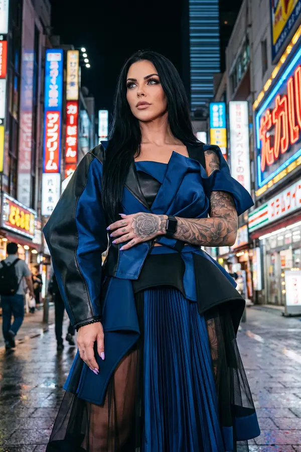 Woman in a navy satin and black leather wrap jacket with a pleated navy skirt under black tulle on a neon-lit street at night
