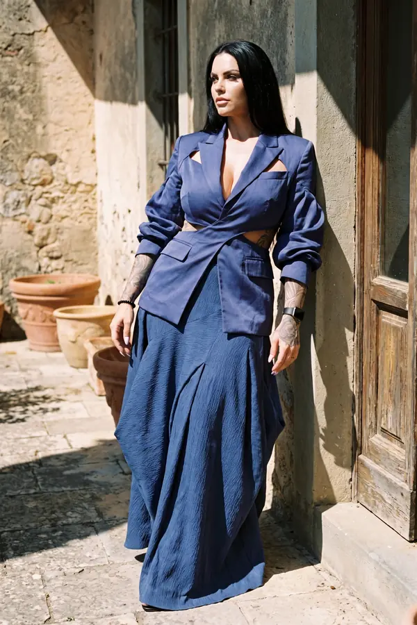 Woman in a navy satin cut-out blazer and a dusty-blue crinkled maxi skirt leaning on a sunlit old stone wall