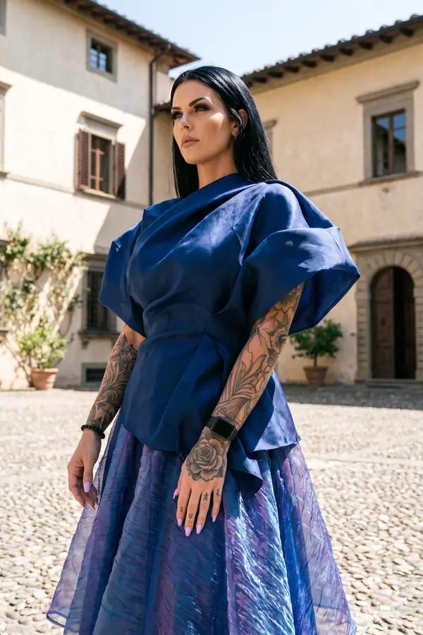 Woman in a navy satin draped top with sculptural cap sleeves and an iridescent organza midi skirt in a stone courtyard