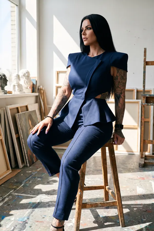 Woman on a wooden stool in a navy sleeveless peplum top with sculpted shoulders and slim trousers in a painter's studio