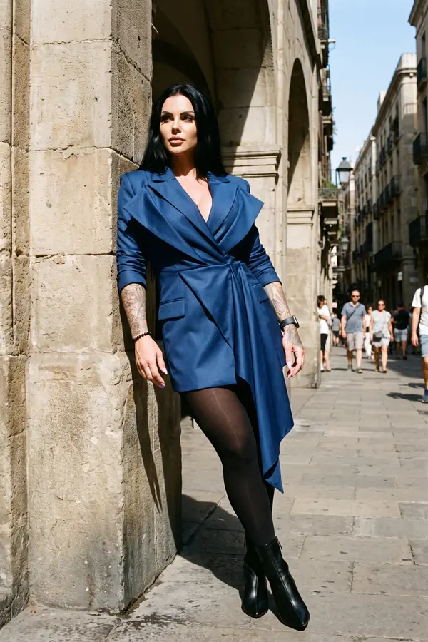 Woman with long black hair in a navy wrap blazer dress, black tights and ankle boots leaning on a stone arcade pillar
