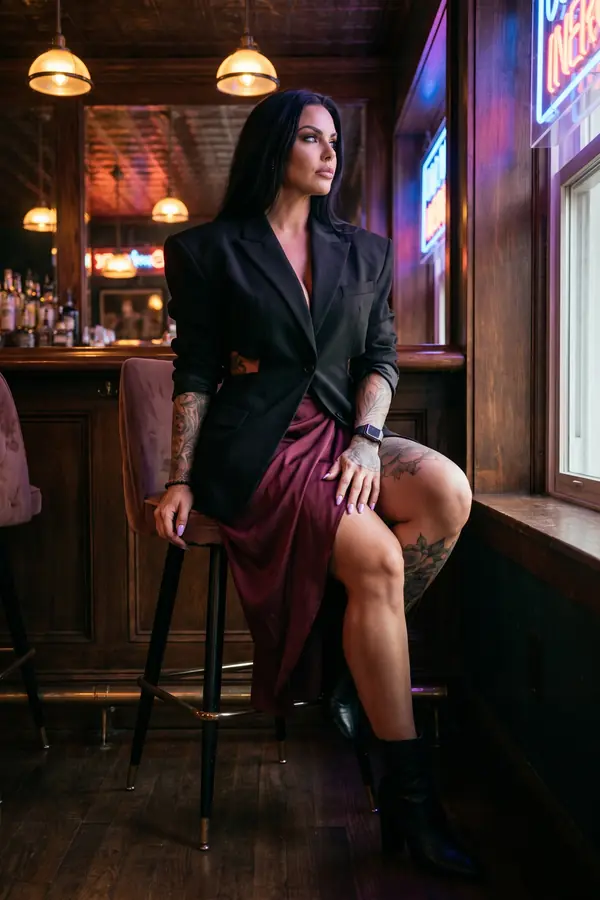 Woman on a bar stool in an oversized black blazer with waist cut-outs, a burgundy satin wrap skirt and black ankle boots