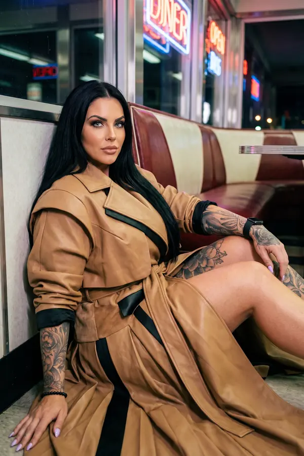 Woman seated on a diner floor against a booth in an oversized camel leather trench with black trim, belted at the waist