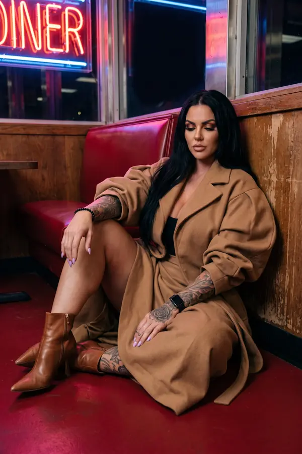 Woman seated on a red diner floor against a booth in an oversized camel wool wrap coat with cognac pointed ankle boots