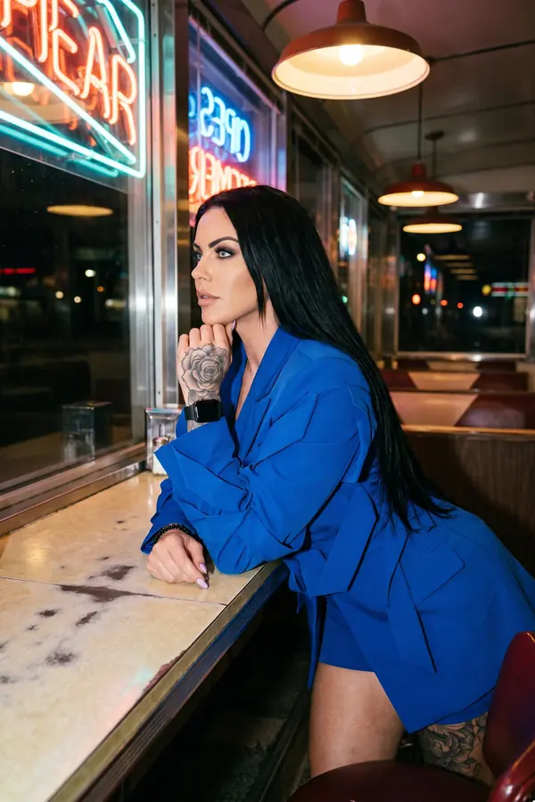 Woman leaning on a diner counter at night in an oversized cobalt blazer dress with layered panel sleeves under neon signs