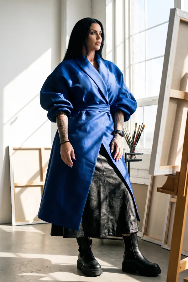Woman in a belted oversized cobalt satin wrap coat over a black leather midi skirt and chunky boots in a sunlit studio