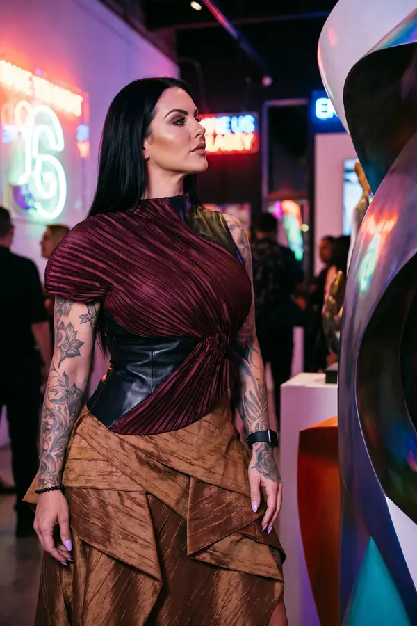 Woman in an oxblood plissé top with a black leather corset panel and a bronze tiered taffeta skirt in a neon-lit gallery